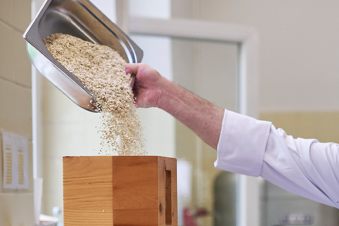 Symbolbild: Küchenpersonal schüttet Hafer in einer hölzerne Mühle.